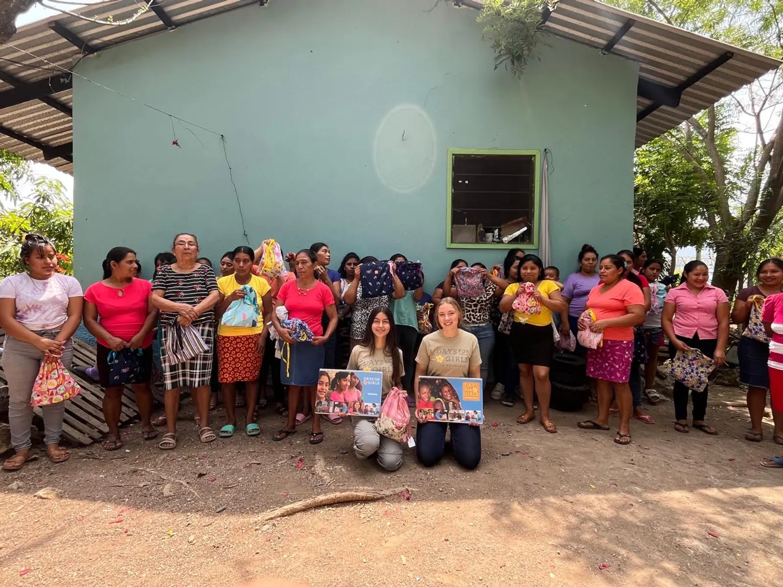 Un grupo de madres y jóvenes con sus kits frente a una casa verde menta en una aldea de Copán; en el centro, dos voluntarias de Days for Girls in Honduras arrodilladas con los calendarios del taller.