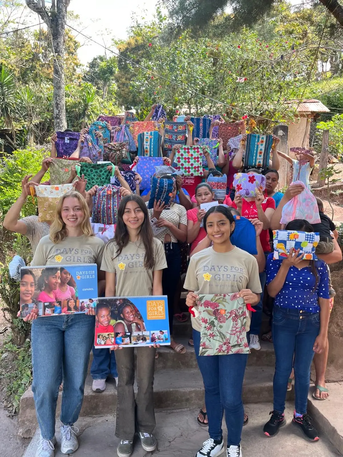 Tres voluntarias de Days for Girls in Honduras, con la camiseta del equipo y los calendarios del taller, en la escalinata de una aldea de Copán; detrás, mujeres y niñas levantan sus kits de tela de colores.