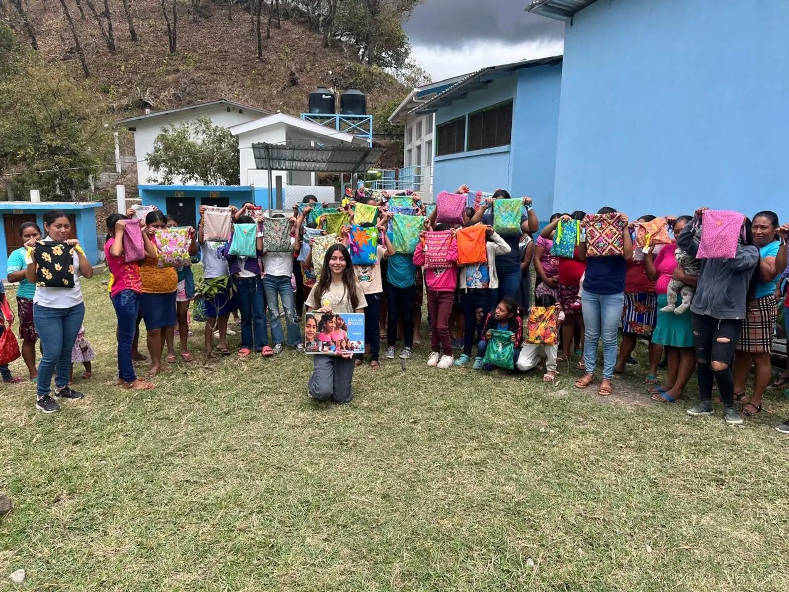 Mujeres y niñas de una aldea de montaña de Copán, en fila frente a una escuela de paredes celestes, cada una con su kit en alto; al frente, arrodillada, una voluntaria con el calendario de Days for Girls.