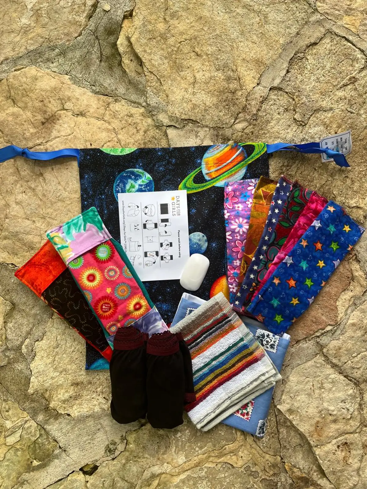 Un kit Days for Girls abierto sobre una piedra: la bolsa de tela con planetas, toallas lavables de colores, dos protectores, ropa interior, jabón, un paño y la hoja de instrucciones.
