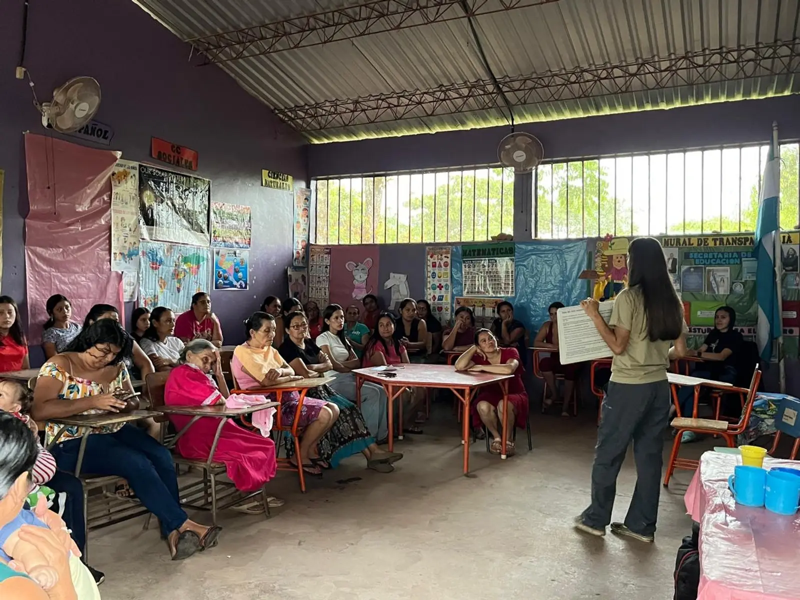 El taller en marcha: en un aula rural de Copán, con carteles escolares en las paredes, mujeres de varias edades sentadas en pupitres escuchan a una voluntaria de Days for Girls que explica de pie con una lámina en la mano.